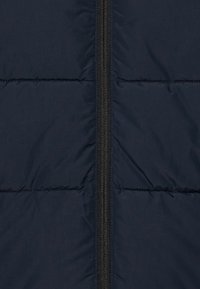 Marinblå vadderad jacka med en central svart dragkedja. Materialet är slätt med sydda horisontella sektioner för isolering.