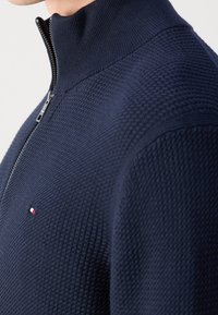 Pull bleu marine avec fermeture éclair, tissu texturé, col montant et petit logo sur la poitrine. Comprend une fermeture éclair argentée.