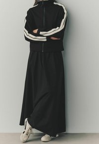 Chaqueta negra con cremallera y rayas blancas, falda maxi negra a juego y zapatillas crema con detalles negros. Textura de tela suave.