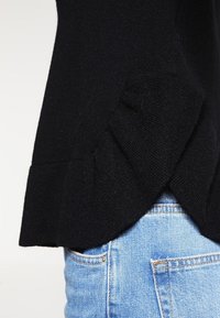 Maglione nero lavorato a maglia con orlo arricciato, che mostra una fine texture. Abbinato a jeans di denim blu chiaro, evidenziando il contrasto di colore.
