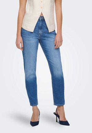 Femme portant un jean en denim bleu, un top beige sans manches boutonné et des chaussures à talons hauts à bride arrière foncées, debout sur fond blanc.
