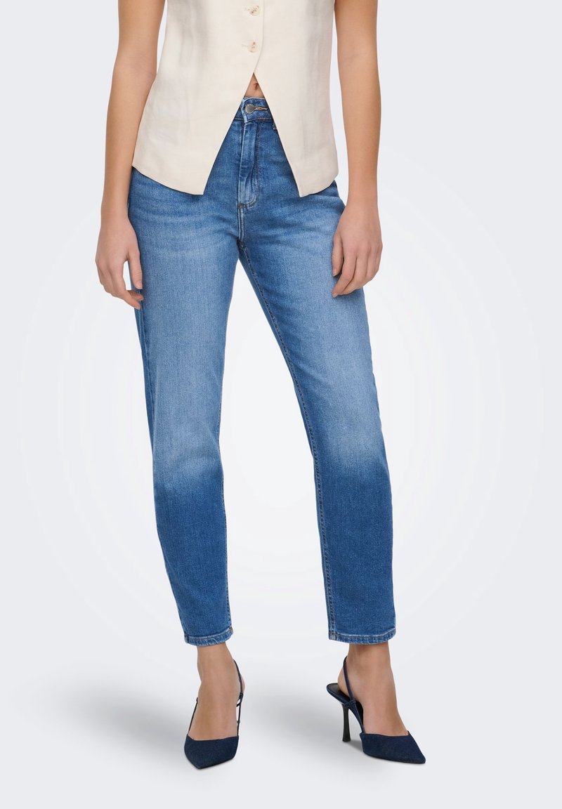 Femme portant un jean en denim bleu, un top beige sans manches boutonné et des chaussures à talons hauts à bride arrière foncées, debout sur fond blanc.