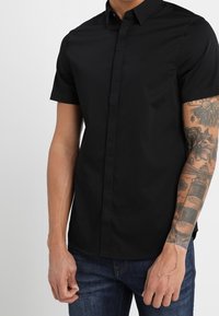 Kortärmad svart skjorta med knappar, tillverkad av en slät tyg, med slim fit och en diskret logotyp på bröstet. Buren med blå jeans.