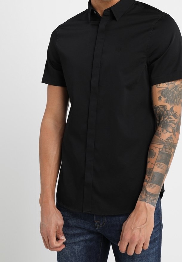 Kortärmad svart skjorta med knappar, tillverkad av en slät tyg, med slim fit och en diskret logotyp på bröstet. Buren med blå jeans.