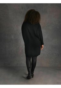 Abito maglione nero a coste con maniche lunghe, vestibilità leggermente oversize e colletto rotondo. Indossato sopra leggings neri e stivali alla caviglia.