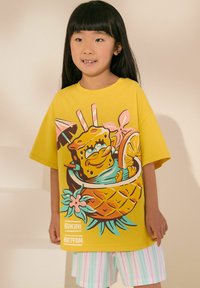 Gul kortærmet t-shirt med et legende cartoonprint af en karakter i en ananas. Kombinationen er med stribede pastelfarvede shorts.