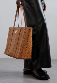 Tan leather tote bag with a patterned surface featuring diamonds and logos, complemented by long handles and a structured silhouette.