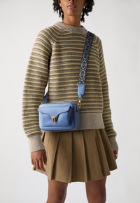 Borsa a tracolla blu in pelle con hardware dorato, superficie testurizzata e tracolla decorata. Indossata con un maglione a righe e una gonna plissettata.