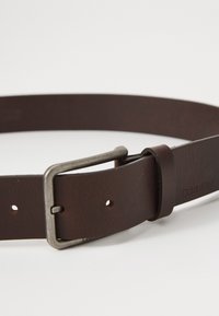 Curea din piele maro cu o cataramă metalică rectangulară. Prezintă detalii de cusătură și mai multe găuri pentru ajustare. Logo-ul gravat este vizibil.