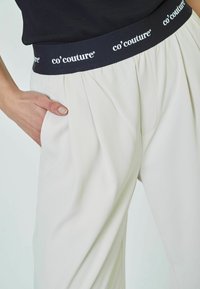 White pleated trousers with a black elastic waistband featuring "co' couture" branding. Side pockets are visible. Smooth fabric texture.