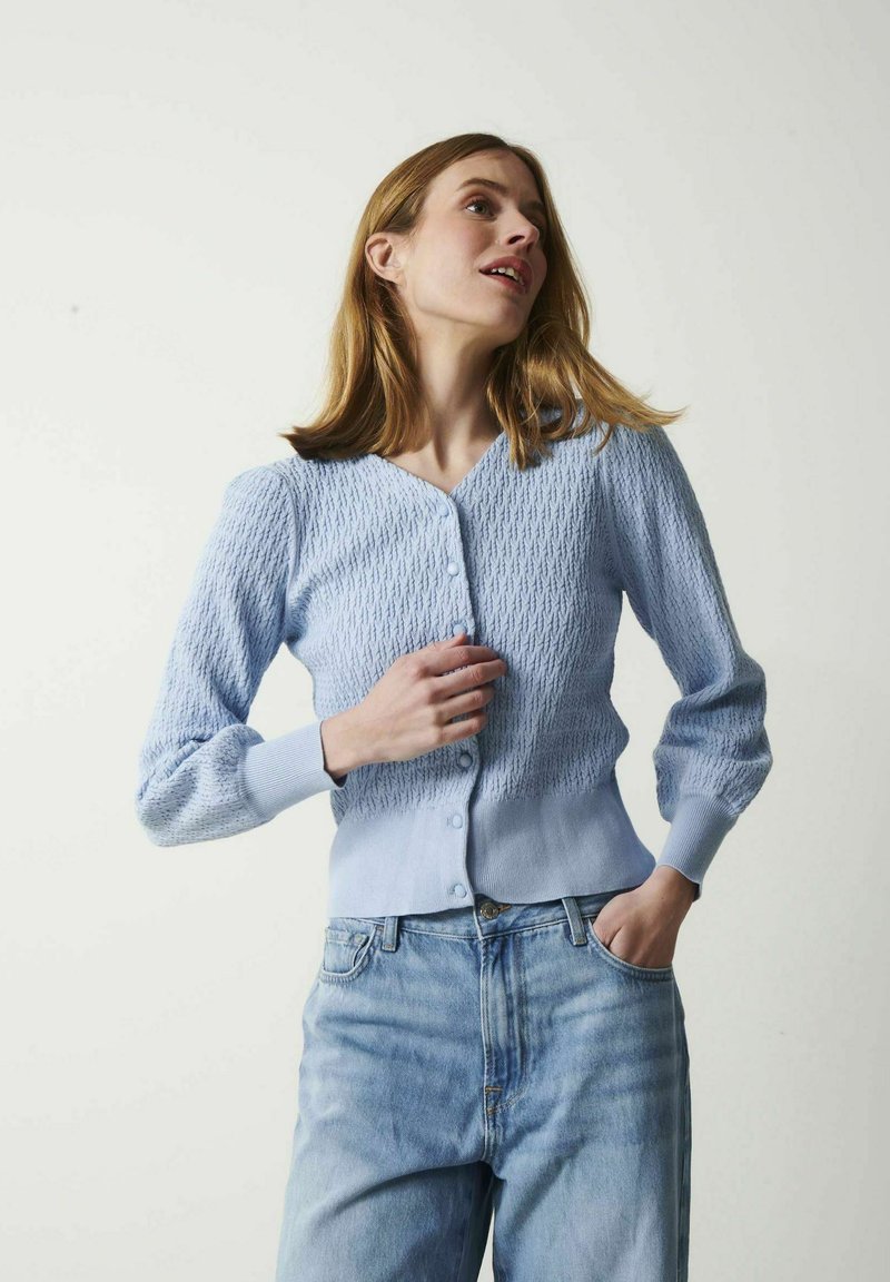 Ung kvinde iført en lyseblå, struktureret cardigan og blå jeans, poserer med den ene hånd i lommen og kigger opad.