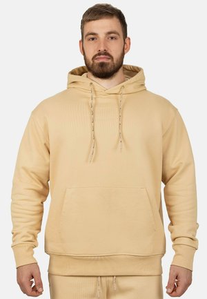 Bärtiger Mann trägt einen beigen Kapuzenpullover mit Fronttasche und passende beige Hose, stehend vor einem einfarbigen Hintergrund.