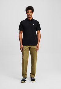 Melna polo krekls ar ribinātu apkakli, logo uz krūtīm, apvienots ar olīvu zaļām biksēm un melniem sporta apaviem ar dzelteniem akcentiem.