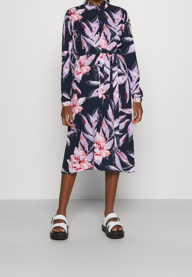 Personne portant une robe midi bleu marine à imprimé floral rose et violet, manches longues, taille ceinturée et sandales blanches à boucle.