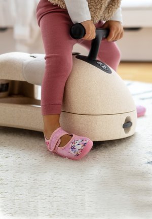Roze kinderschoenen met bloemenpatronen, voorzien van een klittenbandsluiting. Een beige, afgerond object staat op de achtergrond op een lichtgekleurde vloerbedekking.