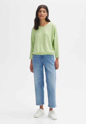 Hellgrünes, geripptes Langarmshirt mit Rundhalsausschnitt und gerafftem Saum, kombiniert mit hellblauen, geraden Jeans und weißen Sneakers.