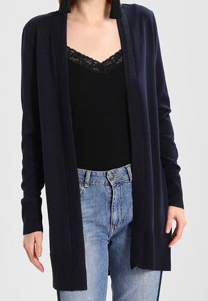 Femme portant un cardigan bleu marine ouvert, un caraco noir orné de dentelle et un jean taille haute bleu clair, sur un fond uni.