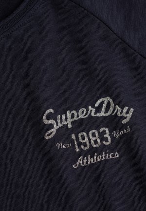 Marineblauw katoenen T-shirt met geribbelde textuur, met zilverkleurige geborduurde tekst: "SuperDry," "New 1983 York" en "Athletics" aan de voorkant.