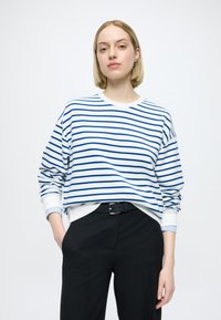 Sweat-shirt rayé en coton blanc et bleu avec un col rond, épaules tombantes, et poignets côtelés, associé à un pantalon noir.