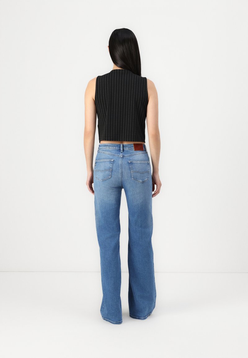 Schwarzes ärmelloses Crop Top mit weißen Streifen, kombiniert mit hellblauen weiten Jeans, die einen ledernen Rückenpatch und Taschen haben.