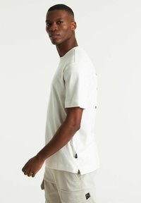 T-shirt a maniche corte, con scollo a girocollo, in cotone beige chiaro. Presenta uno spacco laterale e un piccolo etichetta con il logo sull'orlo. Tessuto liscio.