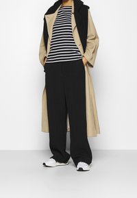 Persona che indossa un trench beige sopra una maglietta a righe bianco e nera, pantaloni larghi neri e sneakers bianche, in piedi con le mani nelle tasche.