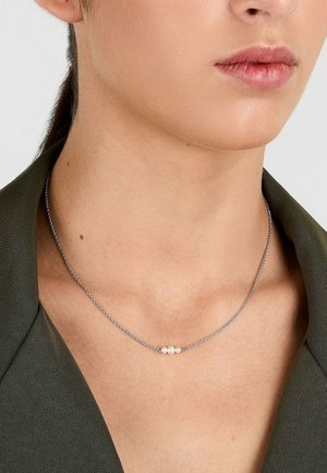 Nahaufnahme einer Person, die eine zarte silberne Kette mit drei kleinen weißen Perlen in der Mitte trägt, gekleidet in ein dunkelgrünes Oberteil.