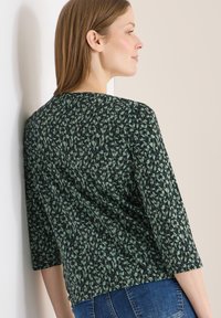 Femme portant un haut à motifs vert foncé et noir avec des manches trois-quarts, vue de profil sur un fond de mur clair.