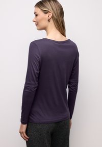 Long-sleeve purple top in soft fabric with a rounded neck, slight stretch, and visible stitching details. Worn with textured grey pants.