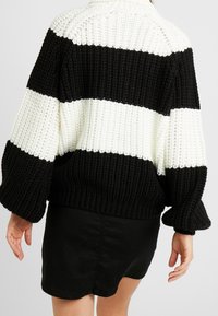 Sweter w dzianinę z czarnymi i białymi poziomymi paskami, grubą teksturą, wysokim kołnierzem i szerokimi rękawami. Noszony na czarnej spódnicy.