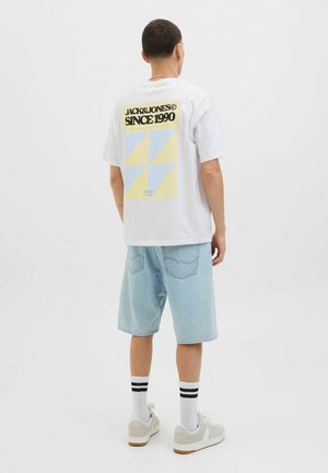 T-shirt blanche à manches courtes avec impression graphique en jaune et bleu, associée à un short en jean bleu clair et des baskets grises avec des accents blancs.