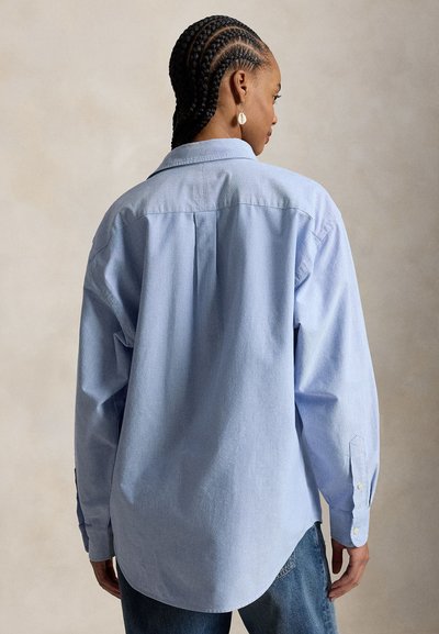 Frau mit geflochtenem Haar, die ein hellblaues Langarmshirt und Jeans trägt, von hinten vor neutralem Hintergrund gezeigt.