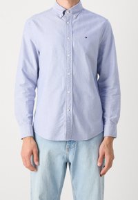 Man die een lichtblauw overhemd met knopen draagt, met een klein logo op de borst en lichte blauwe spijkerbroek, staand tegen een effen achtergrond.