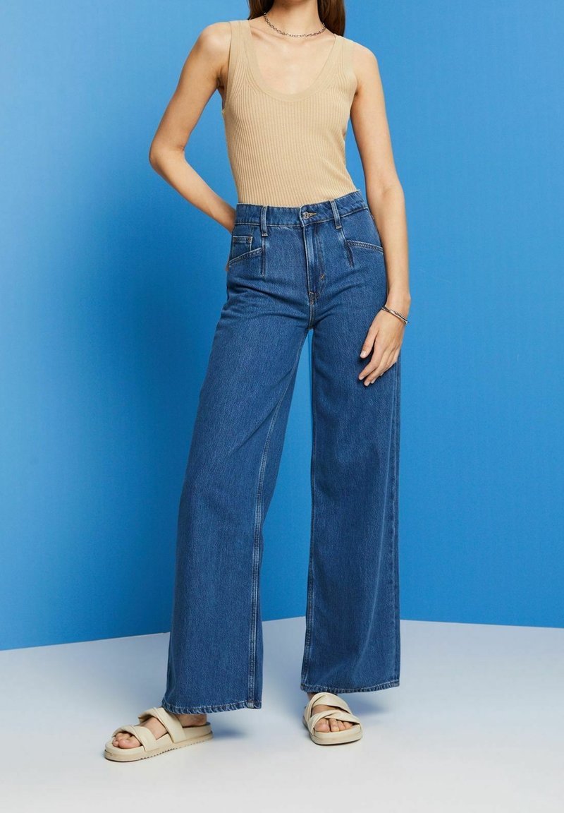 Jeans z wysokim stanem i szerokimi nogawkami w ciemnoniebieskim dżinsie, z zapięciem na guzik, dwoma przednimi kieszeniami i luźnym krojem, w zestawie z beżowym topem na ramiączkach z prążkowanej tkaniny.