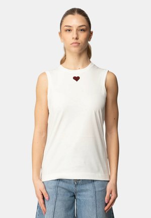 Joven mujer mirando al frente, vestida con una blusa blanca sin mangas con un pequeño corazón rojo bordado en el pecho y jeans azul claro.