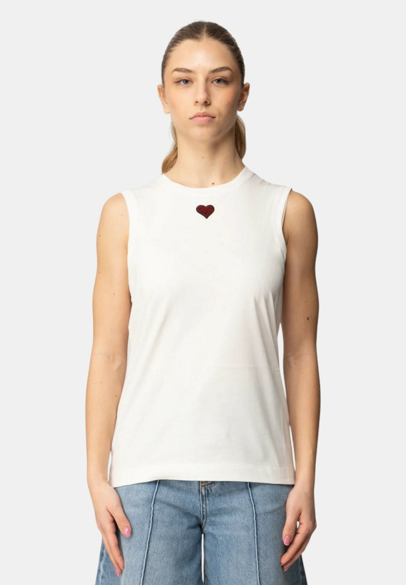 Joven mujer mirando al frente, vestida con una blusa blanca sin mangas con un pequeño corazón rojo bordado en el pecho y jeans azul claro.