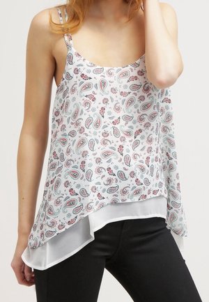 Kvinde iført en hvid ærmeløs top med paisleymønster og sorte bukser, med hånden i håret, vist fra skuldre til hofter.