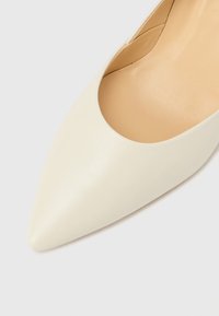 Biely lodičky s vysokým podpätkom a špicatou špičkou z textúrovanej kože. Má hladkú vnútornú podšívku bez viditeľných kovových častí alebo ozdôb.