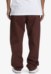 Weite braune Hosen aus robustem Stoff, mit weitem Bein, glatter Textur und lässiger Passform, kombiniert mit weißem Schuhwerk.