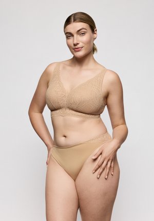 Vrouw met lichte huid en blond haar, gekleed in een beige kanten beha en bijpassend ondergoed, staat zelfverzekerd tegen een effen grijze achtergrond.