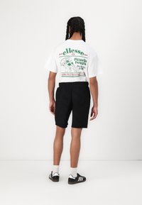 Una camiseta blanca con un diseño gráfico que presenta pizza y texto en verde y rojo. Pantalones cortos negros con un patrón texturizado. Zapatos negros con rayas blancas.