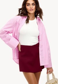 Chemise à rayures roses et blanches avec boutons, portée sur un débardeur blanc, associée à une jupe cramoisie. La chemise a des manches larges et une coupe décontractée.