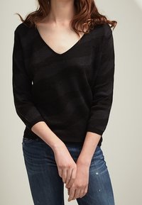 Svart V-ringad tröja med strukturerade ränder, 3/4 ärmar och en lös passform, kombinerad med blå denimjeans.