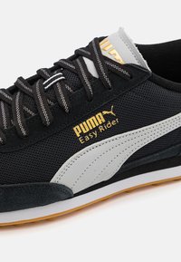 Sapatilha esportiva preta com parte superior em malha, detalhes em camurça cinza, logótipo em dourado "PUMA Easy Rider" e sola de borracha com um padrão texturizado.