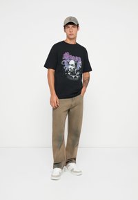 Sort grafisk t-shirt med et trykt design med lilla tekst og et portræt. Beige baggy bukser, grå sneakers med hvide detaljer og en kasket.
