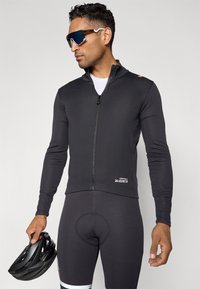 Schwarze Radjacke mit durchgehendem Reißverschluss, langen Ärmeln und körpernahem Schnitt. Kombiniert mit schwarzen gepolsterten Radhosen und einem schwarzen Helm.