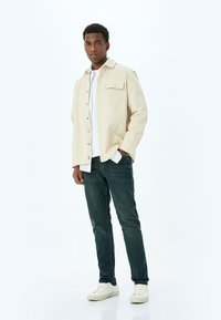 Camisa de botón beige con un bolsillo, superpuesta a una camiseta blanca, combinada con jeans verde oscuro y zapatillas blancas.