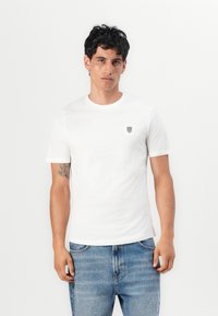 Weißes Baumwoll-T-Shirt mit kurzen Ärmeln, rundem Halsausschnitt und einem kleinen schwarzen Logo auf der linken Brust. Getragen mit blauen Jeans.