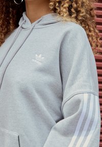 Sweat à capuche gris en tissu doux, avec un petit logo Adidas blanc sur la poitrine et trois bandes blanches sur les manches. Conception avec capuche.