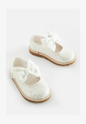 Zapatos de ballet blancos brillantes con suela de goma, que presentan un gran lazo de cinta blanca en la parte delantera y sutiles detalles de costura a lo largo del borde.